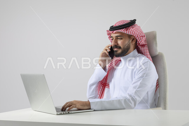 الرد والاجابة على الاتصالات الهاتفية عبر الهاتف المحمول، بورتريه مقرب لرجل عربي خليجي سعودي يرتدي الشماغ والثوب التقليدي يعمل على الحاسب المحمول، دمج التقنية والتكنولوجيا بالحياة العملية، خلفية بيضاء