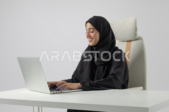  مهن ووظائف نسائية مكتبية سعودية، ادارة وانجاز المهام باستخدام جهاز تقني حديث ومتطور, بورتريه مقرب إمرأة عربية خليجية سعودية مبتسمة ترتدي الحجاب والعباءة تعمل على الحاسوب المحمول, ايماءات وتعابير السرور والسعادة، خلفية بيضاء