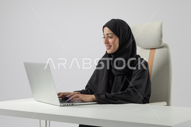  مهن ووظائف نسائية مكتبية سعودية، ادارة وانجاز المهام باستخدام جهاز تقني حديث ومتطور, بورتريه مقرب إمرأة عربية خليجية سعودية مبتسمة ترتدي الحجاب والعباءة تعمل على الحاسوب المحمول, ايماءات وتعابير السرور والسعادة، خلفية بيضاء
