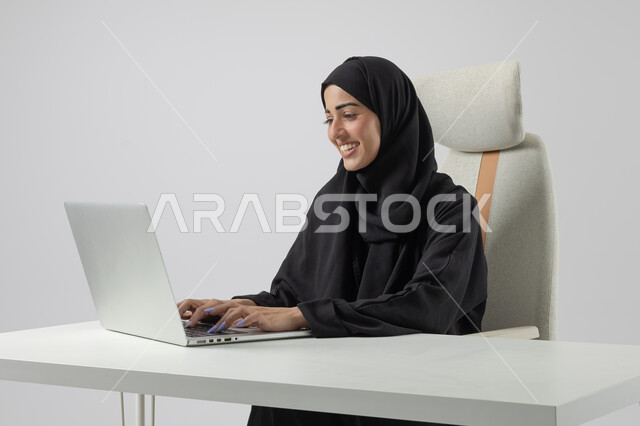  مهن ووظائف نسائية مكتبية سعودية، ادارة وانجاز المهام باستخدام جهاز تقني حديث ومتطور, بورتريه مقرب إمرأة عربية خليجية سعودية مبتسمة ترتدي الحجاب والعباءة تعمل على الحاسوب المحمول, ايماءات وتعابير السرور والسعادة، خلفية بيضاء