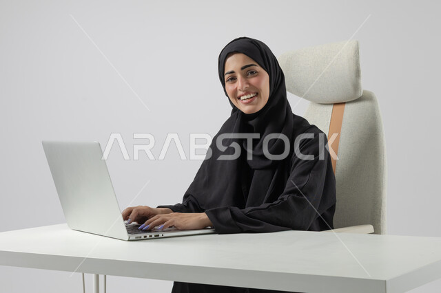  مهن ووظائف نسائية مكتبية سعودية، ادارة وانجاز المهام باستخدام جهاز تقني حديث ومتطور, بورتريه مقرب إمرأة عربية خليجية سعودية مبتسمة ترتدي الحجاب والعباءة تعمل على الحاسوب المحمول, ايماءات وتعابير السرور والسعادة، خلفية بيضاء