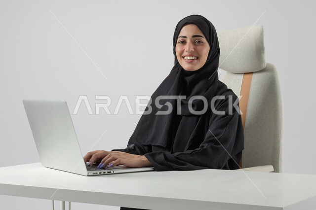  مهن ووظائف نسائية مكتبية سعودية، ادارة وانجاز المهام باستخدام جهاز تقني حديث ومتطور, بورتريه مقرب إمرأة عربية خليجية سعودية مبتسمة ترتدي الحجاب والعباءة تعمل على الحاسوب المحمول, ايماءات وتعابير السرور والسعادة، خلفية بيضاء