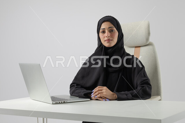  مهن ووظائف نسائية مكتبية سعودية، ادارة وانجاز المهام باستخدام جهاز تقني حديث ومتطور, بورتريه مقرب إمرأة عربية خليجية سعودية مبتسمة ترتدي الحجاب والعباءة تعمل على الحاسوب المحمول, ايماءات وتعابير السرور والسعادة، خلفية بيضاء