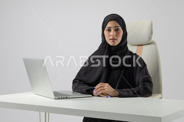  مهن ووظائف نسائية مكتبية سعودية، ادارة وانجاز المهام باستخدام جهاز تقني حديث ومتطور, بورتريه مقرب إمرأة عربية خليجية سعودية مبتسمة ترتدي الحجاب والعباءة تعمل على الحاسوب المحمول, ايماءات وتعابير السرور والسعادة، خلفية بيضاء