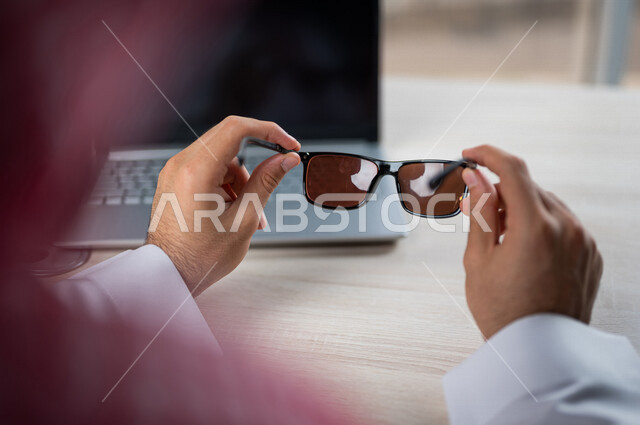 صورة مقربة ليد رجل عربي خليجي سعودي يمسك بيده نظارة شمسية وأماه جهاز الحاسوب المحمول، استخدام تقنيات حديثة، انجاز مهام العمل، اعمال مكتبية