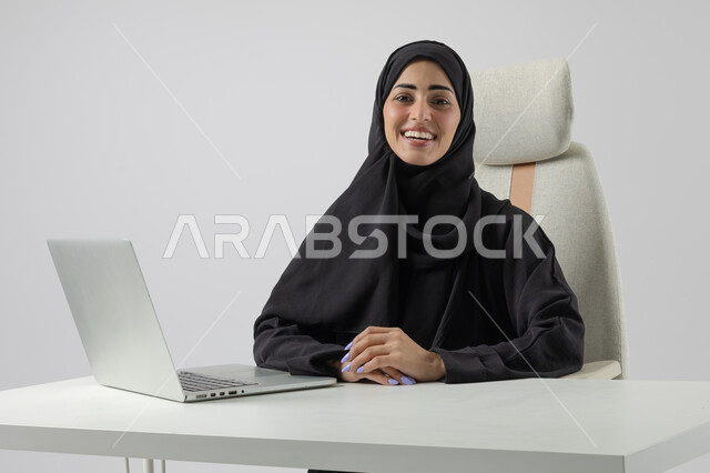 Saudi women's office jobs and professions, managing and completing tasks using a modern and advanced technical device, close-up portrait of a smiling Saudi Arabian Gulf woman wearing a hijab and abaya working on a laptop, gestures and expressions of pleasure and happiness, white background