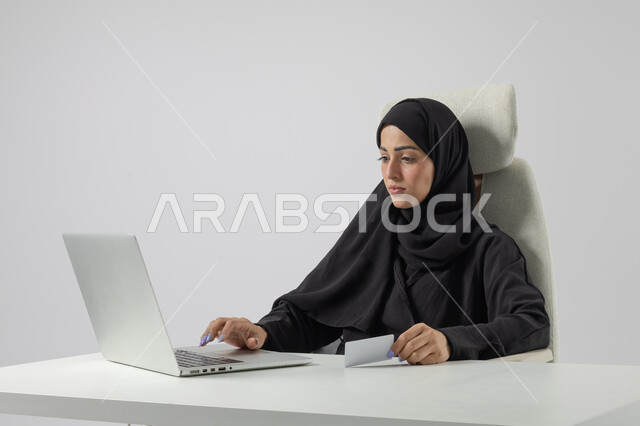  مهن ووظائف نسائية مكتبية سعودية، ادارة وانجاز المهام باستخدام جهاز تقني حديث ومتطور, بورتريه مقرب إمرأة عربية خليجية سعودية مبتسمة ترتدي الحجاب والعباءة تعمل على الحاسوب المحمول, ايماءات وتعابير السرور والسعادة، خلفية بيضاء
