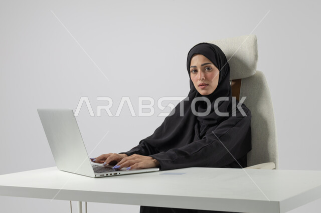  مهن ووظائف نسائية مكتبية سعودية، ادارة وانجاز المهام باستخدام جهاز تقني حديث ومتطور, بورتريه مقرب إمرأة عربية خليجية سعودية مبتسمة ترتدي الحجاب والعباءة تعمل على الحاسوب المحمول, ايماءات وتعابير السرور والسعادة، خلفية بيضاء