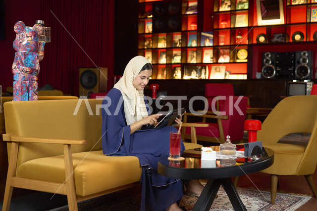 دمج التكنولوجيا المتطورة في الحياة اليوميه، احتساء المشروب المفضل، امراة عربية خليجية اماراتية محجبة ترتدي العباءة تجلس على الأريكة في إحدى المقاهي الراقية تستمع بلحظات الهدوء والراحة ، سيده سعودية تقوم بتصفح مواقع ووسائل التواصل الاجتماعي عبر التابلت