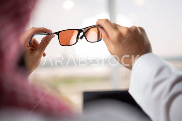 صورة مقربة ليد رجل عربي خليجي سعودي يمسك بيده نظارة شمسية وأماه جهاز الحاسوب المحمول، استخدام تقنيات حديثة، انجاز مهام العمل، اعمال مكتبية