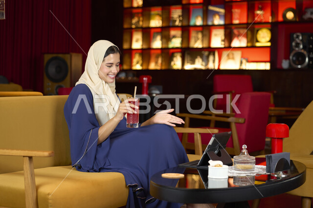 التواصل والتحدث مع الأهل والأصدقاء، احتساء المشروب المفضل، امراة عربية خليجية اماراتية محجبة ترتدي العباءة تجلس على الأريكة في إحدى المقاهي الراقية تستمع بلحظات الهدوء والراحة ، سيده سعودية تقوم باجراء مكالمة فيديو عبر التابلت