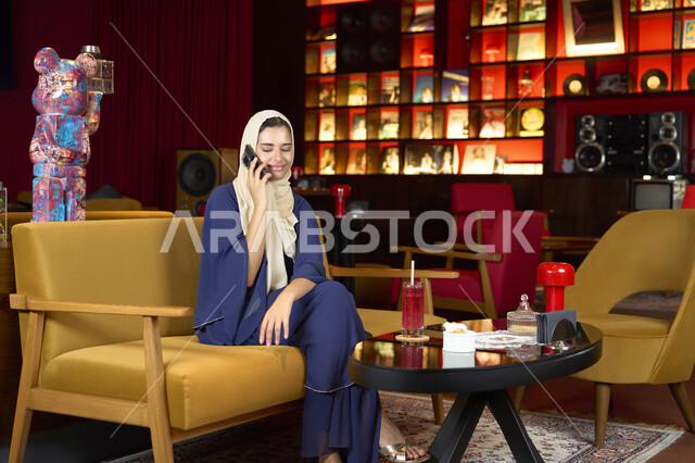 Communicating and checking on friends and relatives, exchanging entertaining conversations via mobile, sipping a favorite drink, a veiled Emirati Gulf Arab woman wearing an abaya sitting on the couch in an upscale cafe enjoying moments of calm and comfort, a Saudi woman making a phone call