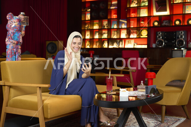 قضاء وقت ممتع عبر الجوال مع الأصحاب، إلقاء التحية والترحيب والسلام، احتساء المشروب المفضل، امراة عربية خليجية اماراتية محجبة ترتدي العباءة تجلس على الأريكة في إحدى المقاهي الراقية تستمع بلحظات الهدوء والراحة ، سيده سعودية تقوم باجراء مكالمة فيديو