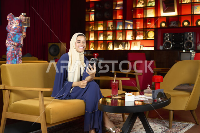 Chatting and socializing, exchanging entertaining conversations via mobile, sipping your favorite drink, a veiled Emirati Gulf Arab woman wearing an abaya sitting on the couch in an upscale cafe enjoying moments of calm and comfort, a Saudi woman making a video call