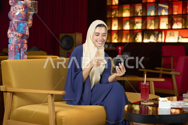 إيماءات الإعجاب والرضا، الدردشة والتواصل مع الاهل والاصدقاء، احتساء المشروب المفضل، امراة عربية خليجية اماراتية محجبة ترتدي العباءة تجلس على الأريكة في إحدى المقاهي الراقية تستمع بلحظات الهدوء والراحة ، سيده سعودية تقوم باجراء مكامله فيديو عبر الهاتف المحمول