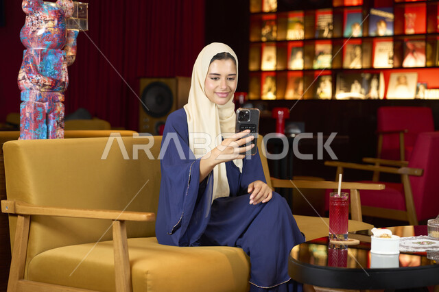 Communicating with friends, having fun outside the home, drinking a favorite drink, a veiled Emirati Gulf Arab woman wearing an abaya sitting on the couch in an upscale cafe enjoying moments of calm and comfort, a Saudi woman making a video call via mobile phone