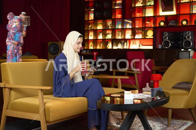 التسوق الالكتروني عبر الانترنت، قضاء أوقات ممتعة ومسلية خارج المنزل، احتساء المشروب المفضل، امراة عربية خليجية اماراتية محجبة ترتدي العباءة تجلس على الأريكة في إحدى المقاهي الراقية تستمع بلحظات الهدوء والراحة ، سيده سعودية تقوم بتصفح الهاتف المحمول