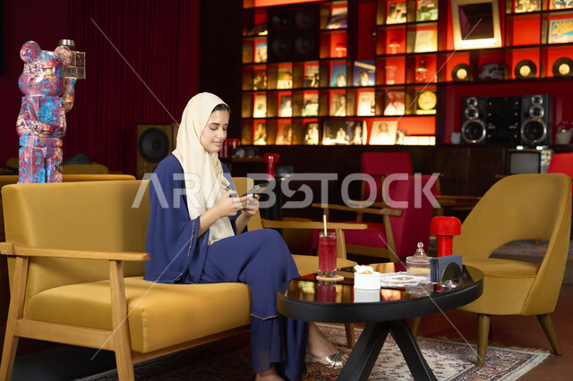 The concept of using technology and modern technology devices, spending fun and entertaining times outside the home, sipping your favorite drink, a veiled Emirati Gulf Arab woman wearing an abaya sitting on the couch in an upscale cafe enjoying moments of calm and comfort, a Saudi woman browsing her mobile phone
