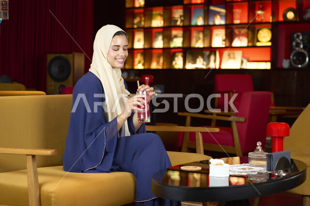 قضاء أوقات ممتعة ومسلية خارج المنزل، الشعور بالانتعاش والفرح, احتساء المشروب المفضل، امراة عربية خليجية اماراتية محجبة ترتدي العباءة تجلس على الأريكة في إحدى المقاهي الراقية تحمل كوب من العصير ، سيده سعودية تستمع بلحظات الهدوء والراحة