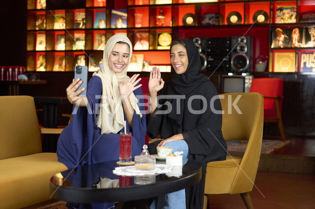 Using modern technology and techniques, documenting happy moments, two Emirati Gulf Arab friends having a drink outside, two Saudi women sitting on the couch in an upscale cafe taking a souvenir photo using their mobile phones