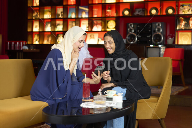 قضاء أوقات ممتعة مع الأصدقاء، النقاش والحوار في الأجواء الهادئة، مشاركة المشروبات الساخنة والقهوة ، صديقتين عربيتين خليجيتين اماراتيتين تقومان بتناول مشروب في الخارج، سيدتان سعوديتان تجلسان على الأريكة في إحدى المقاهي الرقية يقومان بمشاهده الهاتف المحمول