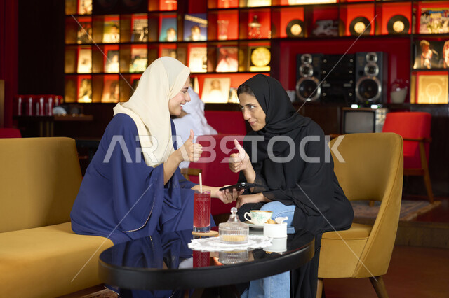 علامة الموافقة و الامتياز، النقاش والحوار في الأجواء الهادئة، مشاركة المشروبات الساخنة والقهوة ، صديقتين عربيتين خليجيتين اماراتيتين تقومان بتناول مشروب في الخارج، سيدتان سعوديتان تجلسان على الأريكة في إحدى المقاهي الرقية يقومان بمشاهده الهاتف المحمول