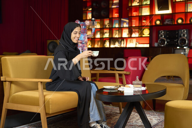 Enjoying hot drinks, recreational youth activities, a smiling Emirati Gulf Arab woman wearing a black abaya sitting on a couch in an upscale cafe, a young Saudi woman drinking a cup of coffee, enjoying the calm atmosphere outside the home