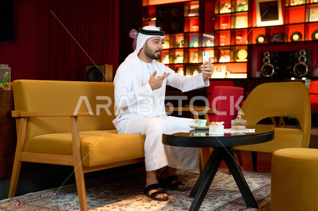 مشاركة الأخبار والأحاديث الجيدة، اجراء مكالمة فيديو مع الاهل والاقارب، الاستمتاع بالاجواء الهادئة خارج المنزل، رجل عربي خليجي اماراتي يرتدي الكندورة والغترة البيضاء يجلس على الأريكة في إحدى المقاهي الراقية يمسك الهاتف المحمول بايماءات الاندماج