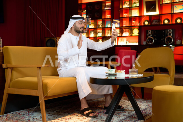 اجراء مكالمة فيديو مع الاهل والاقارب، التواصل مع الاصدقاء عبر الهاتف المحمول، الاستمتاع بالاجواء الهادئة خارج المنزل، رجل عربي خليجي اماراتي يرتدي الكندورة والغترة البيضاء يجلس على الأريكة في إحدى المقاهي الراقية يمسك الهاتف المحمول ،التسلية والترفيه في العطلة