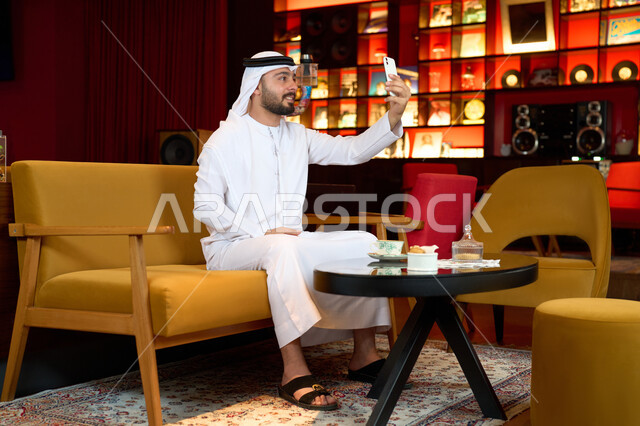 التواصل مع الاصدقاء عبر الهاتف المحمول، اجراء مكالمة فيديو مع الاهل والاقارب، الاستمتاع بالاجواء الهادئة خارج المنزل، رجل عربي خليجي اماراتي يرتدي الكندورة والغترة البيضاء يجلس على الأريكة في إحدى المقاهي الراقية يمسك الهاتف المحمول ،التسلية والترفيه في العطلة