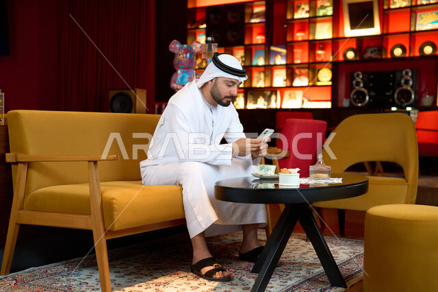 الاستمتاع بالاجواء الهادئة خارج المنزل، رجل عربي خليجي اماراتي يرتدي الكندورة والغترة البيضاء يجلس على الأريكة في إحدى المقاهي الراقية يمسك الهاتف المحمول ،التسلية والترفيه في العطلة، تصفح مواقع التواصل الاجتماعي عبر الانترنت