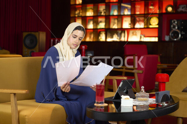 اجواء هادئه خارج المنزل ، الاستمتاع بشرب العصير، ايماءات الاندماج و التركيز، مقاهي في  الامارات العربية، امراة عربيه خليجيه اماراتية محجبه ترتدي العباءة و الحجاب تحمل بيديها اوراق بيضاء، شابه سعودية تقوم بانجاز مهام العمل عبر جهاز التابلت