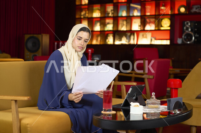 الاستمتاع بشرب العصير، ايماءات الاندماج و التركيز، مقاهي في  الامارات العربية، امراة عربيه خليجيه اماراتية محجبه ترتدي العباءة و الحجاب تحمل بيديها اوراق بيضاء، شابه سعودية تقوم بانجاز مهام العمل عبر جهاز التابلت