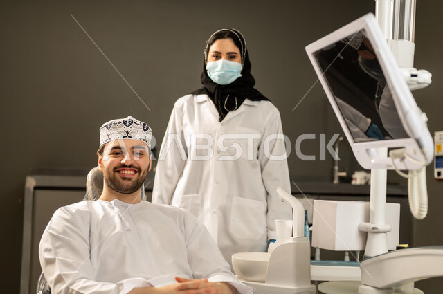 Expressions of happiness and pleasure, providing medical assistance to patients, close-up of a Saudi Arabian Gulf female doctor wearing gloves and a medical mask examining a patient's teeth, oral and dental surgery, a young Saudi Arabian Gulf male wearing traditional clothing sitting in a dental clinic and opening his mouth to diagnose a disease, a young Omani male sitting on a treatment chair.