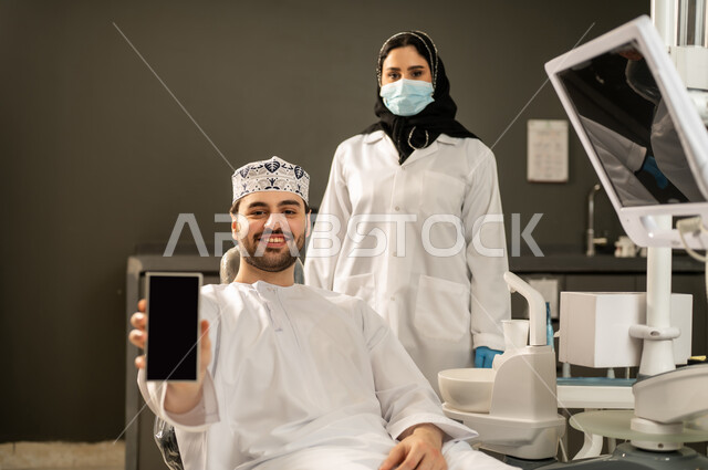 عرض شاشة سوداء فارغة عبر الجوال ,شاب عربي خليجي سعودي يرتدي الزي التقليدي يجلس في العيادة السنية ويفتح فمه لتشخيص المرض, صورة مقربة من الخلف لطبيبة عربية خليجيه سعودية ترتدي القفازات والكمامة الطبية تقوم بفحص أسنان المريض .