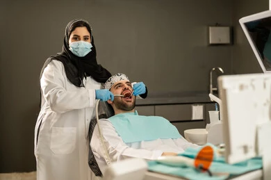 صورة مقربة ليد طبيبة عربية خليجيه سعودية ترتدي القفازات والكمامة الطبية تقوم بفحص أسنان المريض ،إجراء الفحوصات الطبيه ،تقديم الخدمات الصحية في المراكز التخصصية، جراحة الفم والأسنان، شاب عربي خليجي سعودي يرتدي الزي التقليدي يجلس في العيادة السنية ويفتح فمه لتشخيص المرض, شاب عماني يجلس على كرسي العلاج .