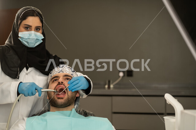 إجراء الفحوصات الطبيه ،تقديم الخدمات الصحية في المراكز التخصصية، جراحة الفم والأسنان، شاب عربي خليجي سعودي يرتدي الزي التقليدي يجلس في العيادة السنية ويفتح فمه لتشخيص المرض, صورة مقربة ليد طبيبة عربية خليجيه سعودية ترتدي القفازات والكمامة الطبية تقوم بفحص أسنان المريض ، شاب عماني يجلس على كرسي العلاج .