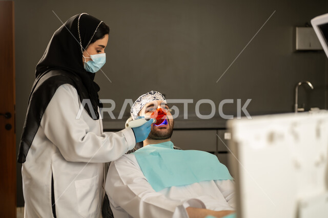 تقديم الخدمات الصحية في المراكز التخصصية، إجراء الفحوصات الطبيه ، جراحة الفم والأسنان، شاب عربي خليجي سعودي يرتدي الزي التقليدي يجلس في العيادة السنية ويفتح فمه لتشخيص المرض, صورة مقربة ليد طبيبة عربية خليجيه سعودية ترتدي القفازات والكمامة الطبية تقوم بفحص أسنان المريض ، شاب عماني يجلس على كرسي العلاج .