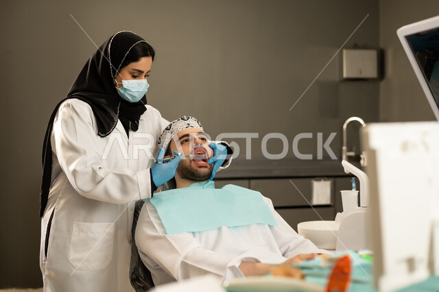 تقديم الخدمات الصحية في المراكز التخصصية ،الأساليب العلاجية المتبعة للعناية بالفم ، شاب عربي خليجي سعودي يرتدي الزي التقليدي يجلس في العيادة السنية ويفتح فمه لتشخيص المرض ، شاب عماني على كرسي العلاج ، صورة مقربة لطبيبة عربية خليجيه سعودية ترتدي القفازات والكمامة الطبية تقوم بفحص أسنان المريض .