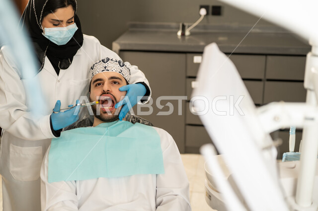 الأساليب العلاجية المتبعة للعناية بالفم ، شاب عربي خليجي سعودي يرتدي الزي التقليدي يجلس في العيادة السنية ويفتح فمه لتشخيص المرض ، شاب عماني على كرسي العلاج ، صورة مقربة لطبيبة عربية خليجيه سعودية ترتدي القفازات والكمامة الطبية تقوم بفحص أسنان المريض .