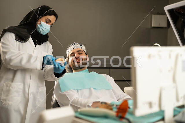 تقديم الخدمات الصحية في المراكز التخصصية ، شاب عربي خليجي سعودي يرتدي الزي التقليدي يجلس في العيادة السنية ويفتح فمه لتشخيص المرض ، شاب عماني على كرسي العلاج ، صورة مقربة لطبيبة عربية خليجيه سعودية ترتدي القفازات والكمامة الطبية تقوم بفحص أسنان المريض .