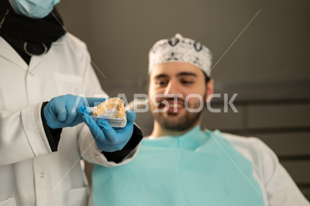 استخدام الادوات اللازمة لإجراء الفحوصات الطبيه , معالجة التسوس و النخر, شاب عربي خليجي سعودي يرتدي الزي التقليدي يجلس في العيادة السنية ويفتح فمه لتشخيص المرض, صورة مقربة لطبيبة عربية خليجيه سعودية ترتدي القفازات والكمامة الطبية تقوم بفحص أسنان المريض ، شاب عماني على كرسي العلاج .