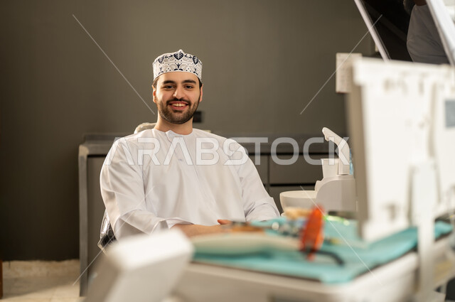 تقديم الخدمات والاستشارات الطبية للمراجعين، النظر الى الكاميرا بايماءات السعادة ، مفهوم الطب والرعاية الصحية ، تدوين معلومات عن حالة المريض، طبيب عماني يرتدي الدشداشة والكمة، مهن ووظائف طبية ، الاهتمام والعناية بالمرضي .