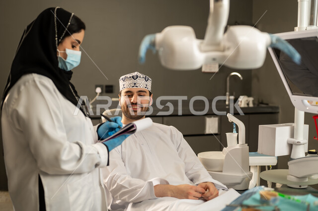 تقديم الخدمات والاستشارات الطبية للمراجعين، مفهوم الطب والرعاية الصحية ،بورتريه لطبيبة عربية خليجية سعودية ترتدي المعطف الطبي تقوم بتدوين معلومات عن حالة المريض، شاب عماني يجلس على كرسي العلاج ، مهن ووظائف نسائية .