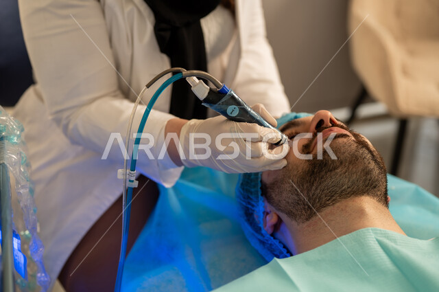 استخدام مكونات تجميلية ، جهاز تنظيف المسام، ترطيب وتفتيح البشرة، صورة لأخصائي جلدية يقوم باستخدام اجهزة العناية بالبشرة ، شاب وسيم عربي خليجي سعودي يرتدي الثوب التقليدي، رجل عماني على كرسي العلاج فى عيادات العناية بالبشرة ، مفهوم العناية والإهتمام بنضارة وتحسين البشرة، إخفاء تجاعيد الوجه، مكافحة الشيخوخة المبكرة.