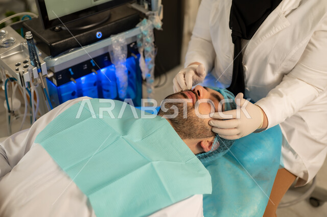 Pore ​​cleansing device, moisturizing and lightening the skin, a picture of a dermatologist applying a mask on the face of a handsome young Arab man from the Gulf of Saudi Arabia wearing traditional clothes, an Omani man on a treatment chair in a skin care center, the concept of caring for and paying attention to the freshness and improvement of the skin, hiding facial wrinkles, fighting premature aging, raising the hand with expressions of caution and fear.