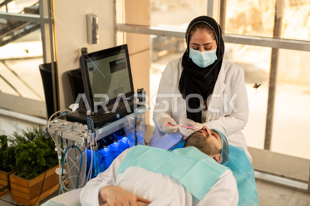 صورة لأخصائي جلدية يقوم بوضع ماسك على وجه شاب وسيم عربي خليجي سعودي يرتدي الثوب التقليدي، رجل عماني على كرسي العلاج فى مركز العناية بالبشرة ، مفهوم العناية والإهتمام بنضارة وتحسين البشرة، إخفاء تجاعيد الوجه، مكافحة الشيخوخة المبكرة، رفع اليد بتعابير الحذر والخوف .
