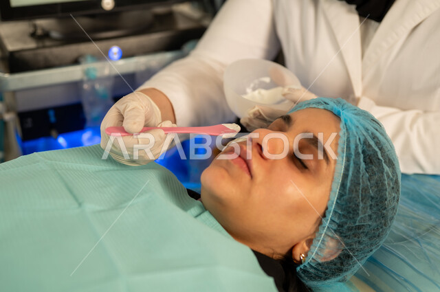 روتين العناية بالبشرة، خبيرة تجميل تقوم بوضع مواد وميك اب على وجه الزبونة، امرأة عربية خليجية سعودية في صالون تجميلي نسائي بايماءات الاسترخاء، استخدام أدوات وفرشاة المكياج، الاهتمام بالجمال والمظهر الخارجي، امرأة عمانية تهتم بمفهوم الأنوثة والنعومة .