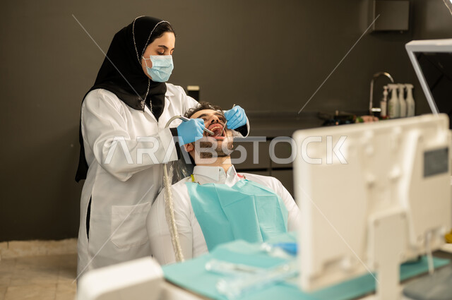 تقديم الخدمات الصحية في المراكز التخصصية ، المعدات اللازمة لإجراء الفحوصات الطبية ، معاينة الحالات المرضية و تشخيصها ، صورة مقربة لطبيبة عربية خليجيه سعودية ترتدي القفازات والكمامة الطبية تقوم بفحص أسنان المريض ، مفهوم الطب والرعاية الصحية ، شاب عربي خليجي سعودي يرتدي الزي التقليدي يجلس في العيادة السنية وينظر الى الكاميرا , شاب عماني يجلس على كرسي العلاج  .