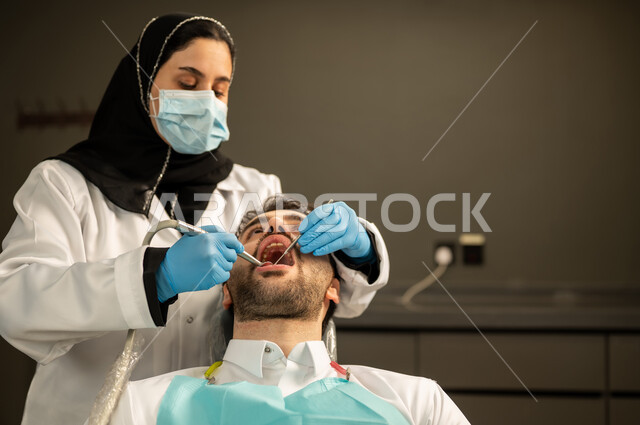 الحفاظ على أسنان المريض , مفهوم الرعاية والعناية بالمرضى ،  صورة مقربة لطبيبة عربية خليجيه سعودية ترتدي القفازات والكمامة الطبية تقوم بفحص أسنان المريض ، مفهوم الطب والرعاية الصحية ، إجراء الفحوصات الدورية للعناية بالصحة الفموية ، شاب عربي خليجي سعودي يرتدي الزي التقليدي يجلس في العيادة السنية وينظر الى الكاميرا , شاب عماني يجلس على كرسي العلاج  .